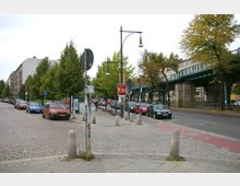 Straßenszene mit parkenden Autos entlang eines gepflasterten Bürgersteigs. Rechts im Bild verläuft eine Hochbahnbrücke aus Stahl, flankiert von Bäumen und Laternen, mit Wohngebäuden im Hintergrund.