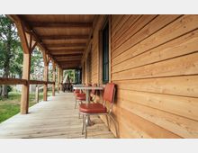 Holzveranda eines Gebäudes mit einer Überdachung aus Holzbalken; an der rechten Seite befindet sich eine mit Holz verkleidete Wand mit Fenstern, links stehen Tische mit roten Stühlen. Im Hintergrund sind Bäume und eine grüne Wiese sichtbar.