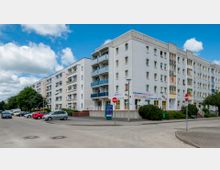 Wohnblock mit mehreren Etagen in moderner Bauweise, weißen Fassaden und Balkonen mit blauen Geländern. Im Erdgeschoss befinden sich Geschäfte, darunter ein Textilservice-Center; davor führt eine Straße mit parkenden Autos entlang.