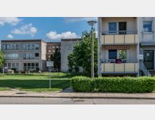 Mehrfamilienhäuser in einer Wohnsiedlung, bestehend aus einem hellen Gebäude mit Balkonen rechts und einem dreigeschossigen Betonbau links. Dazwischen befindet sich eine Grünfläche mit Bäumen, niedrigen Hecken und einem Straßenschild.