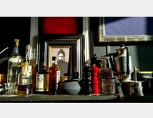 Ein Regal mit verschiedenen Flaschen alkoholischer Getränke, Teekanne, Gewürzmühlen, Glasgefäßen und einer silbernen Kaffeekanne. In der Mitte befindet sich ein gerahmtes Schwarz-Weiß-Porträt einer Frau, umgeben von weiteren dekorativen Elementen.