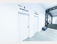 Ein heller, moderner Raum mit weiß gestrichenen Wänden und Türen, die mit Schildern für "3D Body Scan" und "Voice Recording" nummeriert sind. Im Hintergrund ist eine schwarze Metallkonstruktion mit Beleuchtung sowie ein Stativ mit technischer Ausrüstung sichtbar.