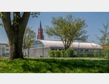 Ein moderner, gewölbter Glasbau mit Metallrahmen steht hinter einem Zaun, umgeben von Bäumen und grünem Rasen. Im Hintergrund ragt der rote Kirchturm einer gotischen Kirche mit spitzem Dach in den blauen Himmel.