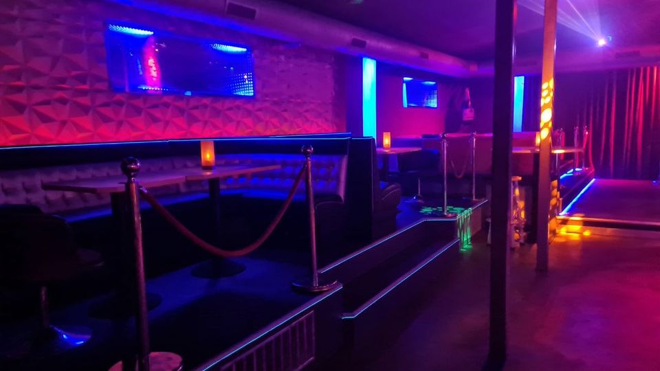 Innenraum eines Nachtclubs mit dunkel gepolsterten Sitznischen und Tischen entlang einer strukturierten Wand, beleuchtet von blauen und violetten Neonstreifen. Rechts befindet sich ein kleiner Bühnenbereich mit vertikalen Stangen und roten Vorhängen, ergänzt durch Akzentlichter und Seilabsperrungen.