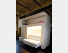 Kompakter Wohnbereich mit integrierter, weißer Schlafcouch in einer modernen Wandkonstruktion. Die Wand aus hellem Material enthält offene Regalfächer mit Büchern, während die indirekte Beleuchtung einen warmen Lichtakzent setzt; sichtbare Ziegeldecke im Hintergrund.