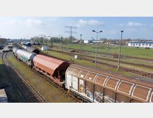 Güterbahnhof mit mehreren Gleisen und abgestellten Waggons, darunter Tank- und Güterwaggons, in einer ländlichen Umgebung mit grünen Wiesen und vereinzelten Gebäuden im Hintergrund. Straßenlaternen und ein Strommast stehen entlang der Gleise.