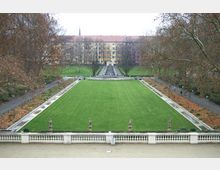 Breiter Park mit einer großen rechteckigen Rasenfläche, gesäumt von Wegen, Bäumen und kleinen Wasserbecken; im Hintergrund befinden sich Stufen, die zu einem Wohngebäude mit gelber Fassade führen. Im Vordergrund ist ein Geländer mit steinernen Figuren sichtbar.