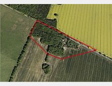 Luftaufnahme einer ländlichen Landschaft mit Feldern und Waldflächen, die von einer roten Linie abgegrenzt wird. Innerhalb der Fläche befinden sich ein Waldstück sowie einige Gebäude am rechten Rand nahe einer Straße.