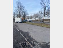 Ein asphaltierter Parkplatz mit vereinzeltem Müll, auf dem ein weißer Anhänger geparkt ist. Im Hintergrund stehen unbelaubte Bäume, parallel zur dahinter verlaufenden Straße mit fahrenden Autos.