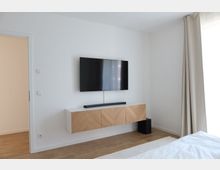 Minimalistisch eingerichtetes Zimmer mit einer weißen Wand, an der ein Flachbildfernseher montiert ist. Darunter befindet sich ein schwebendes Sideboard mit Holzmuster, daneben ein Lautsprecher; der Boden ist aus hellem Holz und rechts hängt ein heller Vorhang.