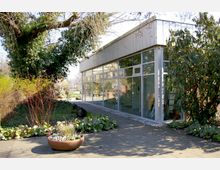 Modernes Gebäude mit Flachdach und großer Glasfassade, umgeben von Bäumen, Sträuchern und einem bepflanzten Blumenbeet. Das Gebäude steht in einer Gartenlandschaft mit gepflastertem Weg.