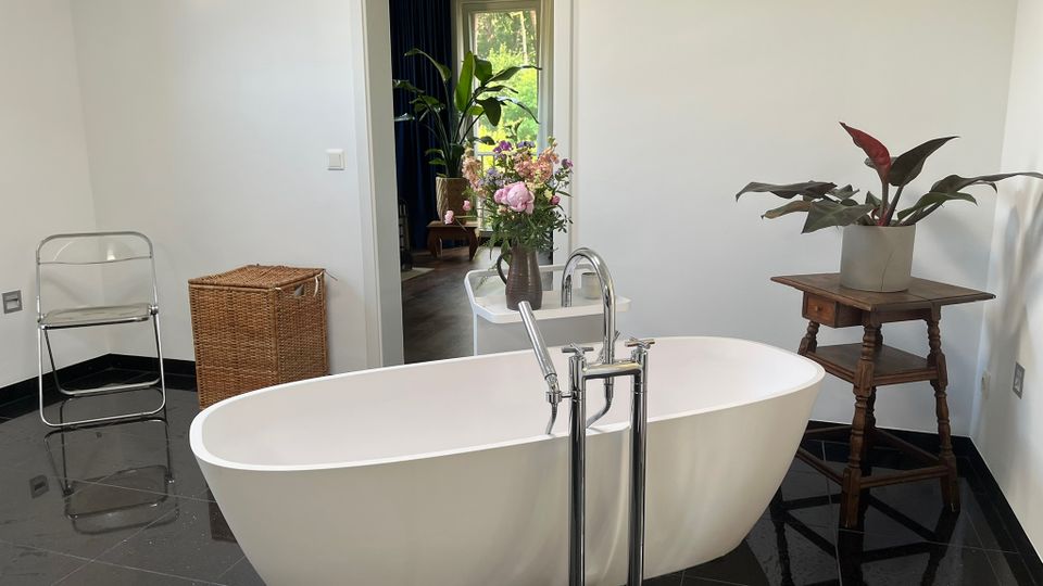 Modernes Badezimmer mit freistehender weißer Badewanne auf glänzendem schwarzen Fliesenboden und bodenstehender Chromarmatur. Neben der Wanne stehen Pflanzen und ein Blumenstrauß, dazu ein kleiner Holztisch; durch eine offene Tür ist ein angrenzender Raum mit Fensterblick ins Grüne zu sehen.