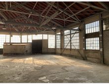 Großer, leerstehender Industriehallenraum mit sichtbarem Stahlträgerdach und roten Holzpaneelen. Die Wände bestehen aus Beton und großen Fenstern mit quadratischen Glasscheiben, durch die Sonnenlicht hereinfällt.