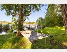 Uferwiese an einem See mit großen Bäumen und einer zwischen zwei Stämmen gespannten Hängematte im Vordergrund. Im Hintergrund führt ein hölzerner Steg mit Geländer ins Wasser, umgeben von Schilf und dichter Ufervegetation.