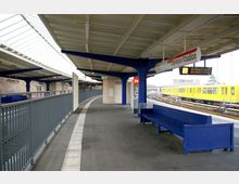Überdachter Bahnsteig eines U-Bahnhofs mit blauen Stahlträgern, einer langen blauen Sitzbank und Geländern. Im Hintergrund fährt ein gelber U-Bahn-Zug auf parallelen Gleisen ein, und ein Schild zeigt den Namen "Olympia-Stadion".
