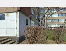 Außenansicht eines mehrstöckigen Gebäudekomplexes mit rau verputzten Fassaden und langen Fensterreihen; links ist ein Seiteneingang mit Stufen und weißer Tür zu sehen. Davor liegen ein kleiner Grünstreifen mit kahlen Hecken und mehreren Bäumen sowie ein schmaler Gehweg entlang des Gebäudes.