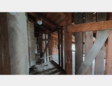 Dachboden eines alten Gebäudes mit freiliegender Holzkonstruktion. Die Wände sind aus grobem Putz und Holzlatten, während der Boden Spuren von Schmutz und Alterung aufweist.