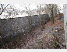 Hinterhof mit kahlen Bäumen und Laub bedecktem Boden, angrenzend an eine hohe Ziegelmauer. Im Hintergrund sind Gebäude erkennbar, und es liegen vereinzelt Gegenstände wie Rohre und ein umgekippter Stuhl herum.