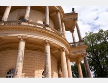 Detailansicht eines historischen Gebäudes mit runden Säulen im korinthischen Stil, einer verzierten Balustrade und gelblicher Fassade. Im Hintergrund sind Bäume und ein bewölkter Himmel sichtbar.