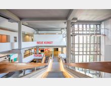 Großer, heller Innenraum eines modernen Museums mit Betonwänden, breiten Treppen und mehreren Etagen. Große Fenster auf der rechten Seite lassen viel Tageslicht herein, und rote Schriftzüge wie "NEUE KUNST" und "FOYER" markieren verschiedene Bereiche.