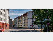 Straßenkreuzung in einer Stadt mit mehrstöckigen Gebäuden, darunter ein weißes Haus mit dem Schriftzug „Jobcenter.“ Verkehrsampeln, ein Straßenschild mit der Aufschrift „Bahnhof“ und Bäume sind sichtbar.