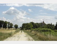 Ländliche Landschaft mit einem unbefestigten Weg, der durch Wiesen und Felder führt. Im Hintergrund ist ein von Bäumen umgebenes Kirchturmgebäude mit rotem Dach sichtbar, unter einem bewölkten Himmel.