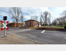 Straßenkreuzung mit einem Bahnübergang, der durch Andreaskreuze und eine Ampel gesichert ist. Im Hintergrund sind Backsteingebäude und eine parkähnliche Grünfläche mit Bäumen sichtbar.