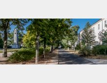 Eine von Bäumen gesäumte Allee mit einem asphaltierten Weg, der zu einem steinernen Gedenkobelisk im Hintergrund führt. Rechts im Bild befindet sich ein historisches, weißes Gebäude mit mehreren Fenstern.