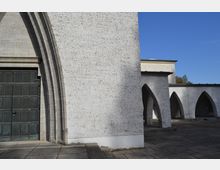 Weiße Fassade eines modernen Gebäudes mit gotischen Elementen, darunter spitzbogige Arkaden und ein großes metallenes Türportal. Der Vorplatz ist gepflastert, und im Hintergrund ist ein blauer Himmel sichtbar.