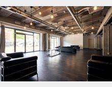 Industrie-inspirierter Innenraum mit offenen Betonwänden und sichtbarer Belüftung an der Decke, ausgestattet mit schwarzen Ledersofas, großen bodentiefen Fenstern und hängenden Glühbirnen. Der Raum hat dunklen Fußboden und bietet viel natürliches Licht durch die Glasfronten, mit Blick auf eine Straße und Gebäude im Hintergrund.