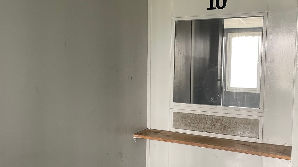 Kleiner, karger Innenraum mit grauen Wänden und abgehängter Decke, an der Stirnwand ein Fenster mit der Nummer „10“ darüber und einer langen, schmalen Ablage darunter. Im Raum steht ein fester, quadratischer Hocker auf einem Metallfuß; der Boden ist dunkel und glatt.