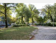 Ein kleiner Stadtpark mit einer Wiese, Bäumen und Pflanzbeeten, umgeben von mehrstöckigen Wohnhäusern. Ein gepflasterter Weg führt durch den Park, auf dem eine Sitzbank mit zwei Personen sichtbar ist.