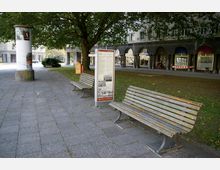 Städtischer Platz mit gepflastertem Weg, Holzbänken, einer Litfaßsäule und einem Infotafelständer, umgeben von Bäumen und Rasenflächen. Im Hintergrund befinden sich Arkaden mit Schaufenstern und Geschäften.