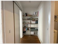 Schmaler Flur mit Holzboden und weißen Wänden, auf der rechten Seite ein offenes Regal mit Büchern, Zeitschriften und dekorativen Gegenständen, links eine weiße Schiebetür.