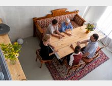 Holzgetäfelter Raum mit einem rechteckigen Holztisch, um den fünf Personen sitzen. Eine gepolsterte Holzbank mit rotem Stoff steht an der Wand, ergänzt durch Holzstühle, eine bunte Blumenvase und einen orientalischen Teppich auf dem Boden.