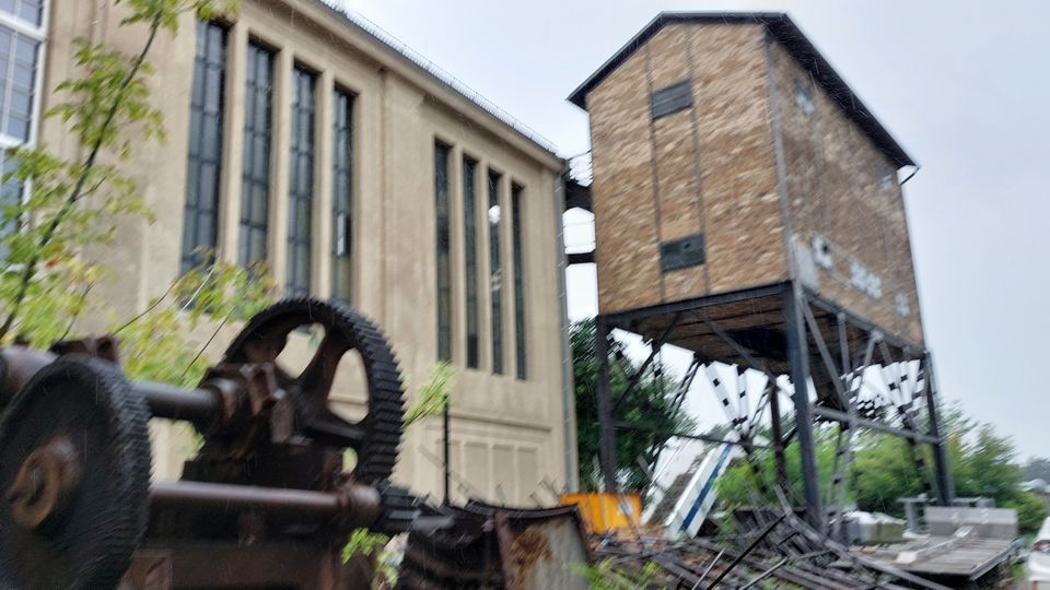 Industriegelände mit einem historischen Fabrikgebäude und einem erhöhten Holzkonstrukt auf Metallstützen. Im Vordergrund sind alte Maschinenteile und Schrott zu sehen, umgeben von vereinzeltem Grün.