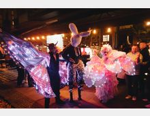 Nächtliche Außenansicht eines überdachten Bar- und Veranstaltungsbereichs mit warmen Lichterketten, Pflasterboden und dem Leuchtschild „BAR 100“. Im Vordergrund sind dekorative, bunt beleuchtete Kostüm- und Flügelelemente zu sehen, im Hintergrund stehen Tische und Eingangsbereiche unter dem Vordach.