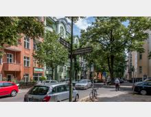 Straßenszene in einer Wohngegend mit mehrstöckigen Gebäuden in verschiedenen Farben (orange und grün) und Balkonen. Im Vordergrund stehen Straßenschilder ("Zimmermannstraße" und "Rothenburgstraße"), geparkte Autos, ein Fahrrad sowie Bäume, die Schatten spenden.