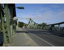 Grüne Stahlbrücke mit geschwungenem Geländer und dekorativen Bögen, die eine Straße mit Fahrbahn und Gehweg über eine Wasserfläche führt; im Hintergrund Bäume und blauer Himmel.