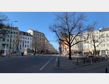 Straßenszene in einer Stadt mit mehrstöckigen Wohnhäusern im Altbaustil auf beiden Seiten, einem breiten Gehweg, kahlen Bäumen und einem zentralen Straßenkreuzungspunkt. Fahrräder und Geschäfte mit Schildern und Schaufenstern sind entlang der Erdgeschosse zu sehen, bei klarem blauem Himmel.