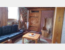 Wohnraum mit rustikalem Design, ausgestattet mit Holzmöbeln, darunter ein Ledersofa, ein kleiner Holztisch und ein geschnitzter Holzstuhl. Die Wände sind aus Holz und Ziegeln, verziert mit runden Holzeinsätzen und Glaselementen, während eine Holztür mit Metallbeschlägen zentral sichtbar ist.