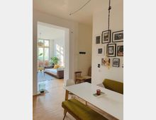 Helles Wohnzimmer mit Esstisch im Vordergrund, einer grünen Polsterbank und mehreren hängenden Lampen. Im Hintergrund ein angrenzender Wohnbereich mit großen Fenstern, einem Sofa und Pflanzen, die den Raum dekorieren.