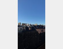 Blick auf die Dächer einer städtischen Wohngegend mit Schornsteinen und Dachfenstern unter einem klaren, blauen Himmel. Im Vordergrund ist ein dunkler Sichtschutz aus Bambusstäben zu sehen.