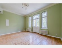 Leerer Wohnraum mit hellgrünen Wänden, hohen Decken mit Stuckrosette und Holzfußboden. Drei hohe Fenster sowie eine verglaste Flügeltür zur Außenseite, darunter Heizkörper; an der Wand ist eine Klimaanlage montiert.