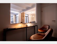 Moderner Wartebereich mit zwei lederbezogenen Stühlen und einem schmalen Holztisch, auf dem zwei kleine Tischlampen und Zeitschriften stehen. Durch ein großes Fenster ist ein Büro mit Backsteinwänden und großen Fenstern sichtbar.