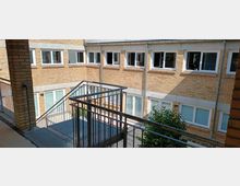 Innenhof eines Gebäudekomplexes mit gelben Backsteinwänden und mehreren weißen Fenstern. Im Vordergrund befindet sich eine Metalltreppe mit Geländer, die in den Hof führt, und ein kleiner Baum ist im unteren Bereich sichtbar.