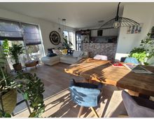 Offenes Wohnzimmer mit moderner, gemütlicher Einrichtung, hellem Holzboden und großen Fenstern mit blickdichtem Rollos. Im Vordergrund ein massiver Holztisch mit Stühlen, im Hintergrund eine Couch, zahlreiche Zimmerpflanzen und eine Küche mit Ziegelwand.