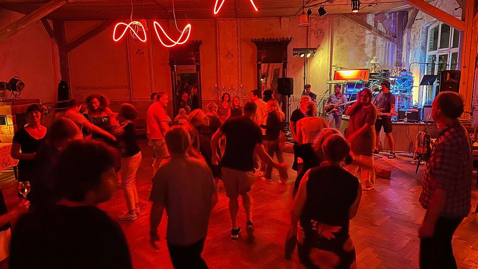 Großer Veranstaltungsraum mit Holzboden und sichtbaren Deckenbalken, beleuchtet von roten Neonröhren und Bühnenscheinwerfern. Rechts steht eine kleine Bühne mit Musikanlage und Instrumenten vor hohen Fenstern und einer teils abgenutzten Wand.