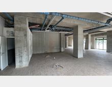 Leerer Rohbau eines Gebäudes mit Betonwänden, freiliegenden Rohrleitungen an der Decke und einem Boden aus grauen Fliesen. Große Fensterfronten lassen Tageslicht in den Raum, und offene Verkabelungen liegen auf dem Boden.