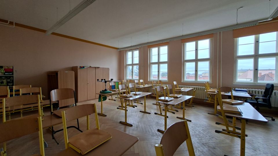 Klassenzimmer mit mehreren Schülertischen und -stühlen, teils umgedreht auf den Tischen, sowie Schränken an der Rückwand. Große, mehrteilige Fenster mit Heizkörpern darunter lassen Tageslicht herein; Holzboden und lange Deckenleuchten prägen den Raum.